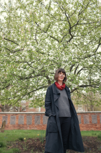 Sarah stands beneath a tree. She wears glasses, a red scarf, and a long black coat. 